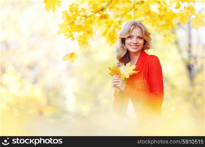 Cheerful young woman sitting on the ground covered with dry autumnal foliage in beautiful park