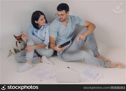 Cheerful young woman uses banking app on her mobile phone, his husband shows figures on calculator, surrounded with papers, have much work, solve economic problems, discuss something together