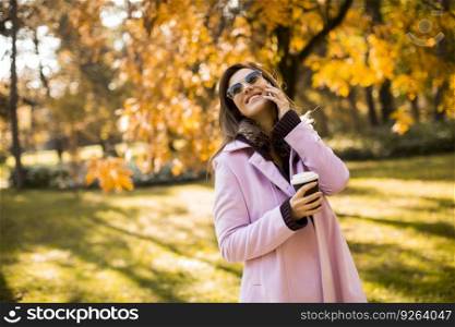 Cheerful young woman wearing pink coat using her phone in the sunny park and drinking take away coffee