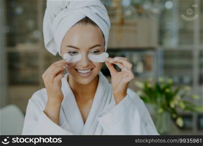 Cheerful young woman with healthy skin takes off beauty patches from under eyes reduces wrinkles or puffiness wears wrapped towel on head and dressing gown smiles toothily has minimal makeup
