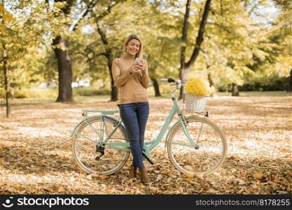 Cheerfull young woman with bicycle using smartphone in autumn park