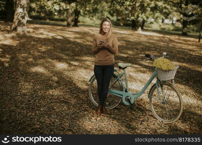 Cheerfull young woman with bicycle using smartphone in autumn park