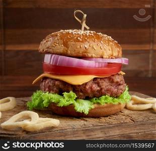 cheeseburger with tomatoes, onions, barbecue cutlet and sesame bun on an old wooden cutting board, brown background. Fast food