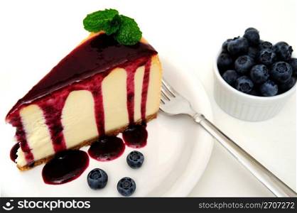 Cheesecake Dessert. Plain Cheesecake with a Blueberry sauce poured over the top with fresh berries on the plate next to the cake and topped with a mint leaf.