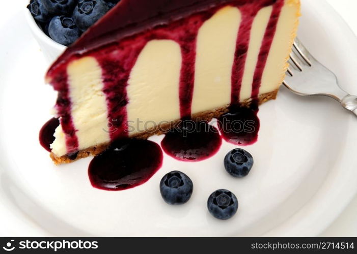 Cheesecake With Blueberry Sauce. Plain Cheesecake with a Blueberry sauce poured over the top with fresh berries on the plate next to the cake and topped with a mint leaf.