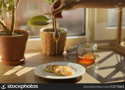 Cheesecakes or syrniki on a plate with honey, in the background a jar with honey and ficus. a woman pouring wooden spoon for honey which flows honey. Close up. selective focus.. Cheesecakes or syrniki on a plate with honey, in the background a jar with honey and ficus. a woman pouring wooden spoon for honey which flows honey. Close up. selective focus