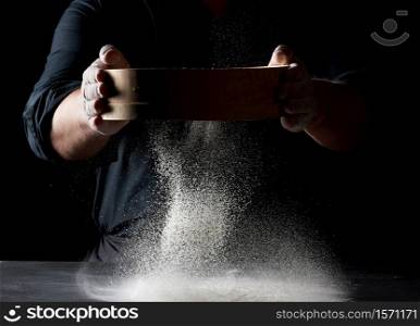 chef a man in a black uniform holds a round wooden sieve in his hands and sifts white wheat flour on a black background, the particles fly in different directions