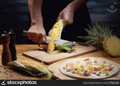 Chef cooking hawaiian pizza, cutting fresh corn. Low key shot, close up of hands, some ingredients around on table.