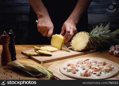 Chef cooking hawaiian pizza, cutting fresh pineapple. Low key shot, close up of hands, some ingredients around on table.