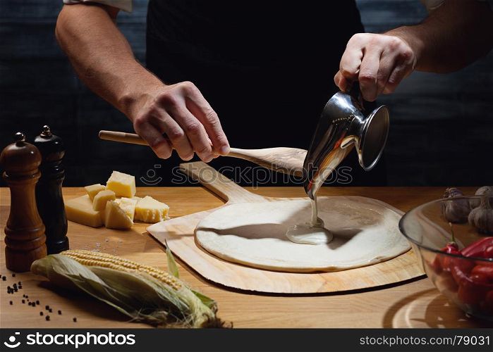 Chef cooking pizza, putting white sauce on pizza base. Low key shot, close up of hands, some ingredients around on table.