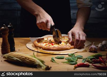 Chef cutting freshly baked hawaiian pizza. Low key shot, close up of hands, some ingredients around on table.
