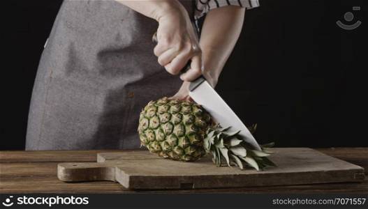 Chef girl in an apron cur juicy tropic fruit pineapple on two parts on a wooden cutting board on a kitchen table on a black background. Motion, 4K UHD video, 3840, 2160p.. Woman chef&rsquo;s hands are cutting fresh natural pineapple on two halves on a wooden board on a kitchen table on a black background. Motion, 4K UHD video, 3840, 2160p.