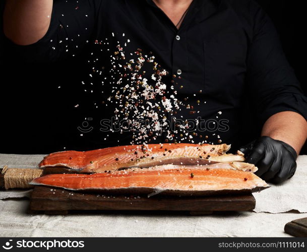 chef in a black shirt and black latex gloves prepares salmon fillet on a wooden cutting board, process of sprinkling with spices and salt, low key