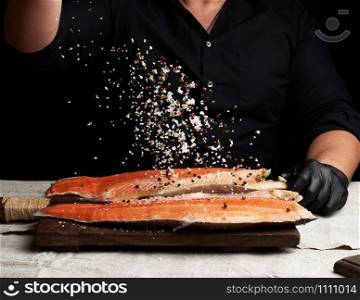 chef in a black shirt and black latex gloves prepares salmon fillet on a wooden cutting board, process of sprinkling with spices and salt, low key
