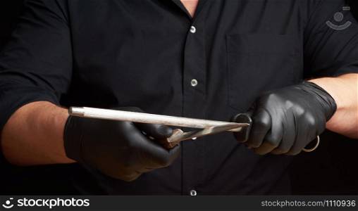 chef in a black shirt and black latex gloves sharpen a kitchen knife on an iron sharpener with a handle above the table, low key, close up