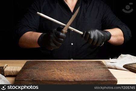 chef in a black shirt and black latex gloves sharpen a kitchen knife on an iron sharpener with a handle above the table, low key