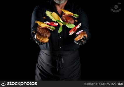 chef in a black shirt, apron and latex black gloves stands on a black background, in his hands flying cheeseburger ingredients: a bun with sesame seeds, cutlet, tomato, lettuce and onion rings, cheese