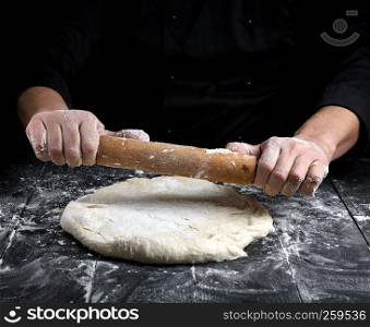 chef in a black tunic rolls a dough for a round pizza on a black table with a wooden rolling pin
