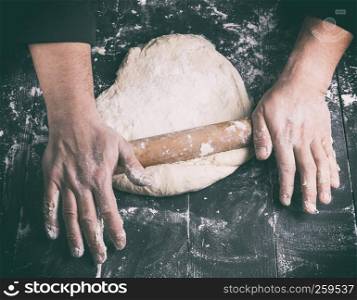 chef in a black tunic rolls a dough for a round pizza, top view