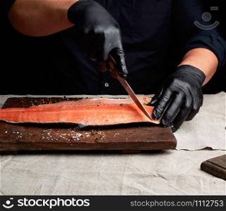 chef in black clothes and black latex gloves cuts fresh salmon filet into pieces, dark key