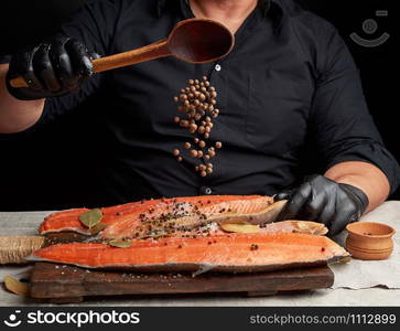 chef in black clothes and black latex gloves pours allspice on a sliced carcass of fresh salmon fish, low key, spices soar in the air