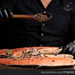 chef in black clothes and black latex gloves pours allspice on a sliced carcass of fresh salmon fish, low key