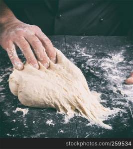 Chef in black jacket, kneads dough from white wheat flour on a black wooden table