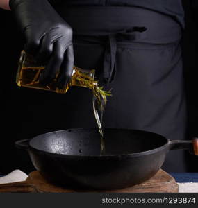 chef in black latex gloves pours olive oil from a transparent bottle into a black cast-iron frying pan, close up