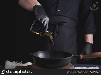 chef in black latex gloves pours olive oil from a transparent bottle into a black cast-iron frying pan, close up