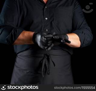 chef in black shirt and apron puts black latex gloves on his hands before preparing food, black background