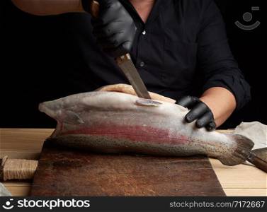 chef in black shirt and black latex gloves slices a whole fresh fish salmon on a vintage brown cutting board, low key