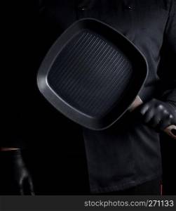 Chef in black uniform and black latex gloves holding an empty square grill pan, black background