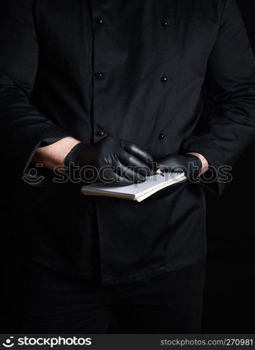 chef in black uniform and latex gloves holding a blank notebook and a black wooden pencil, black background