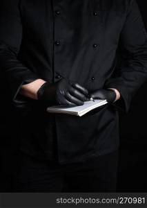 chef in black uniform and latex gloves holding a blank notebook and a black wooden pencil, black background