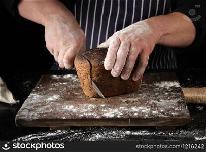 chef in black uniform holds a kitchen knife in his hand and cuts off pieces of bread from a baked brown rye flour loaf on a wooden board