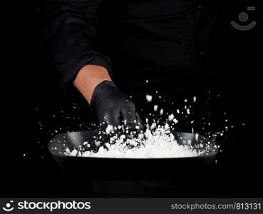 Chef in black uniform holds a pan with white salt and throws up, selective focus, blurred grains of seasoning