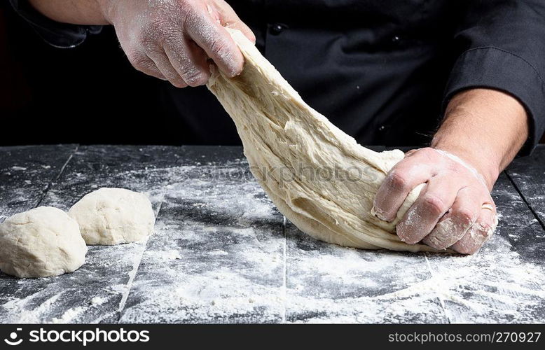 chef in black uniform kneads white wheat flour dough on a black wooden table