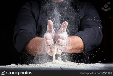 chef in black uniform pours white wheat flour out of his hands on a wooden table, the product flies to the side