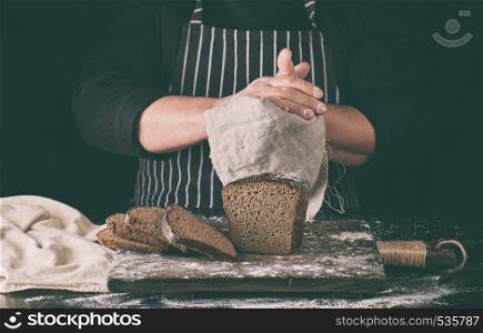 chef in black uniform sliced baked rye bread on a brown wooden board sprinkled with white flour and wipes his hands with a gray bark napkin, black background