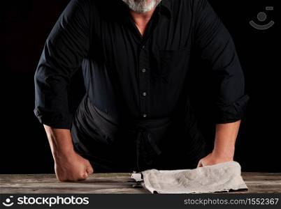 chef man in black uniform bent over a wooden table and rested his fists, black background