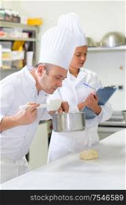 chef smelling contents of saucepan colleague with clipboard in background
