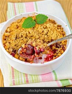 Cherry crumble in a white bowl with a spoon and mint on a napkin on a wooden boards background