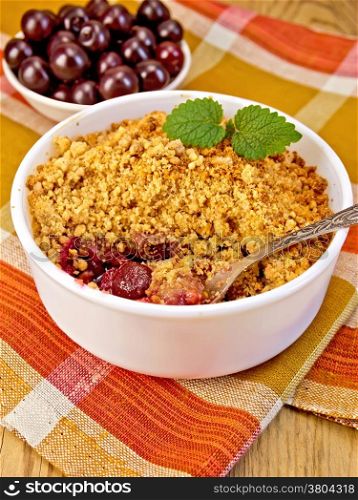 Cherry crumble in a white bowl with a spoon on a napkin, cherries on a background of wooden boards