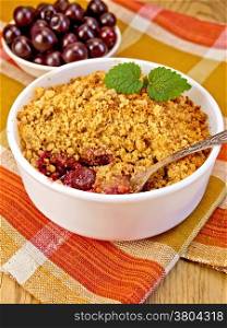 Cherry crumble in a white bowl with a spoon on a napkin, cherries on a background of wooden boards