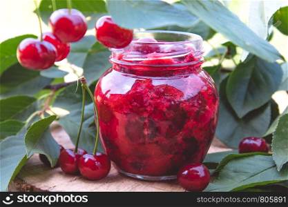 Cherry jam in a glass jar on a background of green leaves on a table of wooden boards. Close-up.. Cherry jam in a glass jar on a background of green leaves on a table of wooden boards.