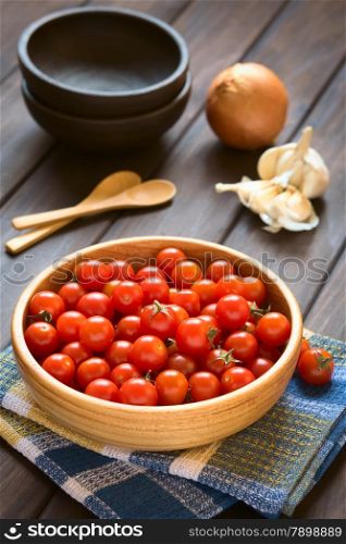 Cherry tomatoes in wooden bowl with garlic, onion, bowls and wooden spoons in the back, photographed on wood with natural light (Selective Focus, Focus one third into the tomatoes)