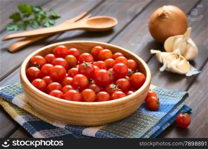 Cherry tomatoes in wooden bowl with garlic, onion, parsley leaves and wooden cutlery in the back, photographed on wood with natural light (Selective Focus, Focus one third into the tomatoes)