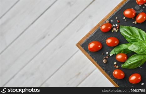 Cherry tomatoes with basil leaves, salt and pepper, layout on a black stone board. Ingredients for making Caprese salad. Copy space on white wooden background, top view