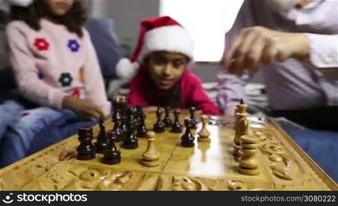 Chess game for clever minds on winter holidays. Diverse family playing chess during christmas at home. Selective focus on players hands moving chess pieces with beautiful smiling mixed race girl in santa hat wathcing match on background. Dolly shot.