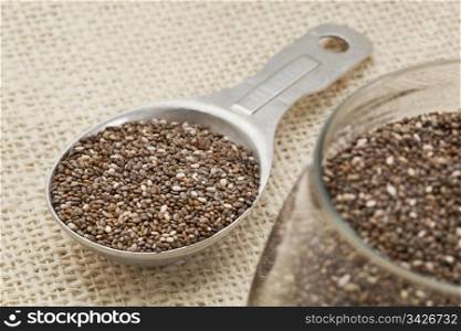 chia seeds in glass jar and on measuring aluminum tablespoon against burlap background, focus on the spoon
